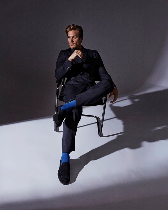 Homme en costume bleu foncé et chaussettes bleu vif, assis sur une chaise.