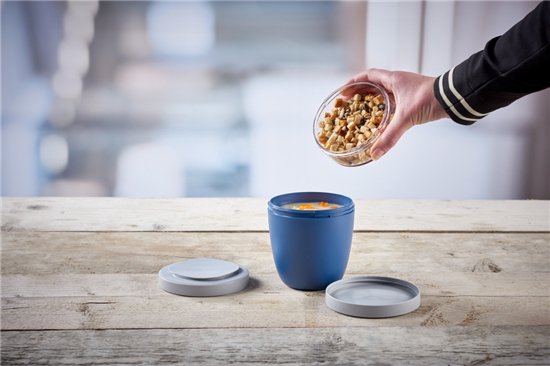 Blauwe lunchpot op tafel, met een hand die walnoten toevoegt uit een glazen potje.