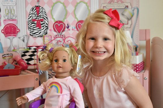 Een meisje met blond haar en een rode strik speelt met een pop met blond haar, beiden in een roze setting.