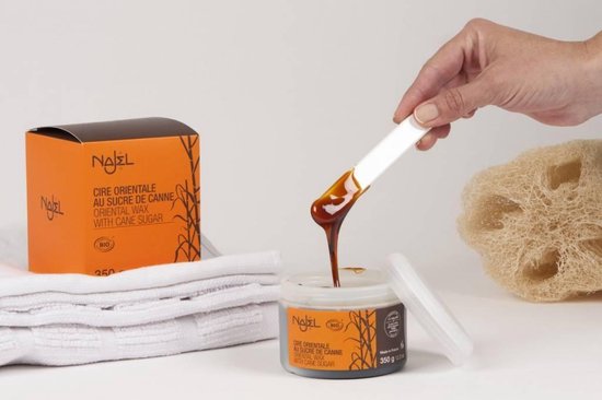 Oranje doosje en potje met Najel Oosterse suikerwax. Een hand houdt een spatel met wax uit het potje. Wit handdoekje en loofah erbij.