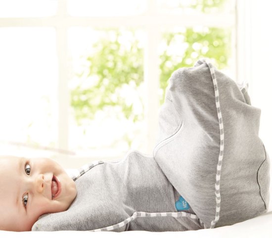 Baby met blauwe ogen ligt in grijze Swaddle UP slaapzak met streepjesrand, met benen omhoog, op witte ondergrond.