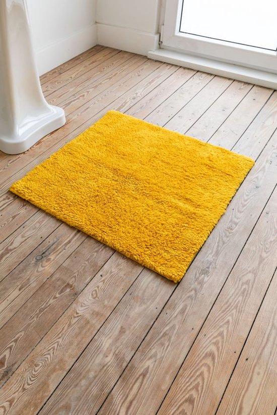 Een heldergele, vierkante badmat van 60x60 cm ligt op een houten vloer naast een witte wastafel.