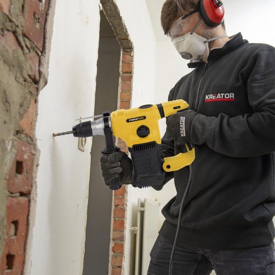Man met veiligheidsuitrusting gebruikt gele klopboormachine om een muur te doorboren. Tekst: '1500W - 5J, 0-4000 slagen/min'.