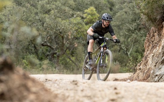 Man op een groene mountainbike met gele accenten, rijdend op een stoffig pad in het bos.