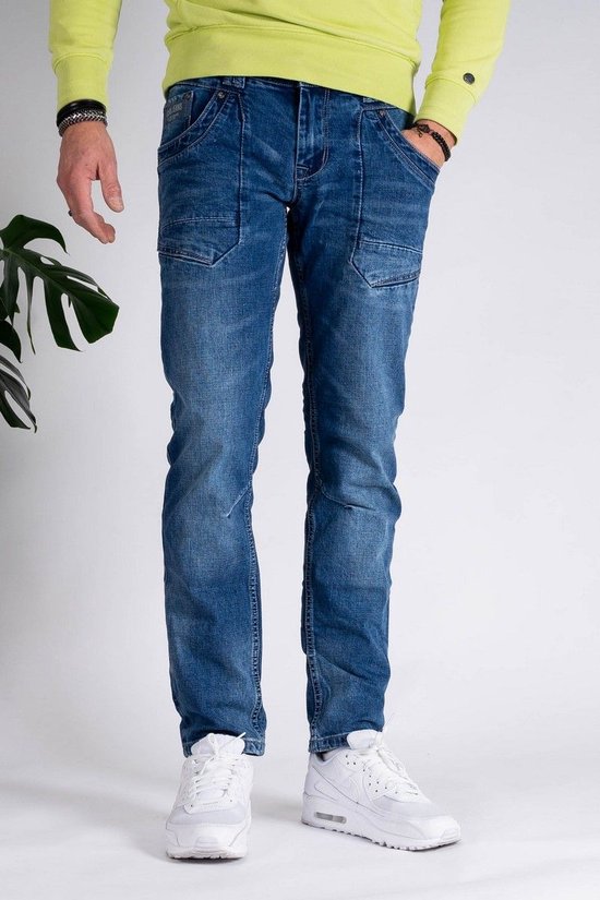 Man in een lichtgroene trui en een stonewashed blauwe jeans. De jeans heeft extra naden op de bovenbenen en witte sneakers.