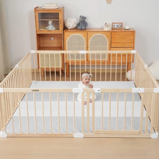 Baby in een ruime houten babybox met witte speelmat en een deur.