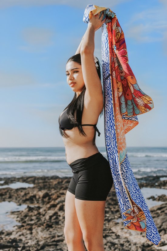 Vrouw met zwarte bikini en korte broek houdt een felgekleurd, bedrukt strandlaken omhoog op een rotsachtige kustlijn.