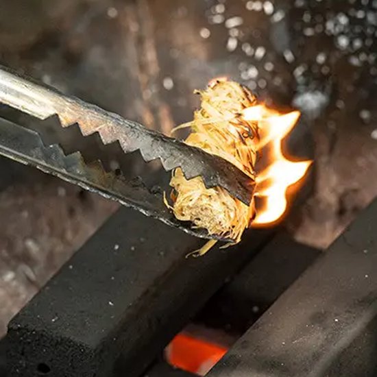 Tang houdt een brandende aanmaakwokkels boven hete kolen.