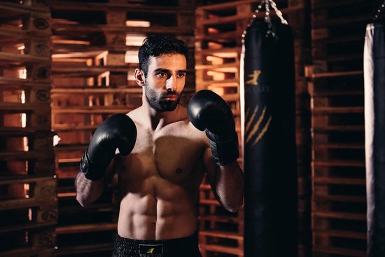 Een gespierde man met zwarte bokshandschoenen, die traint in een gymzaal met houten paletten en bokszakken.