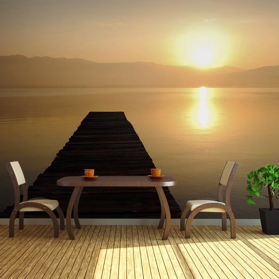 Table avec deux chaises et une plante sur une terrasse en bois avec un papier peint photo d'un ponton au coucher du soleil.