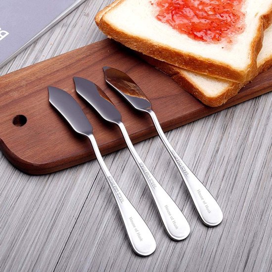 Drie zilverkleurige botermesjes met 'STAINLESS STEEL' en 'House of Husk' inscriptie op een houten plank naast toast met jam.