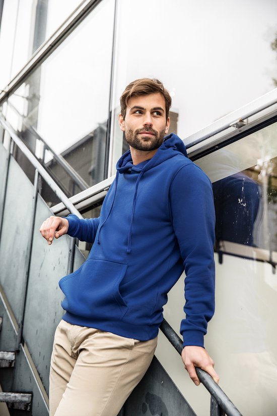 Man met bruin haar en baard in een koningsblauwe hoodie en beige broek, leunend op een trap.
