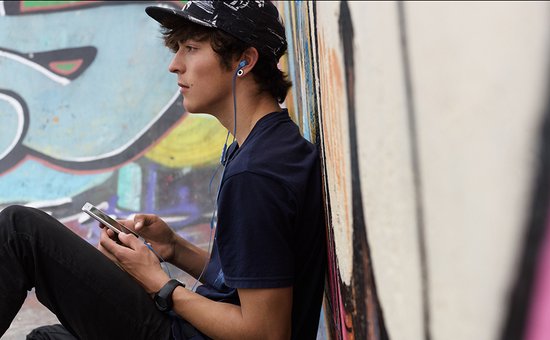 Jongeman met blauwe oordopjes en zwarte pet, luisterend naar muziek via smartphone bij graffiti.