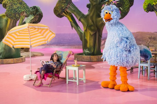 Een grote, pluizige blauwe vogel met oranje poten staat op een roze ondergrond. Een vrouw zit in een ligstoel onder een gele parasol.