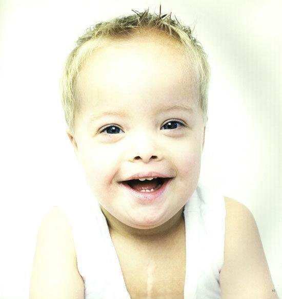 Een jongetje met blond haar en blauwe ogen, draagt een wit hemdje en lacht breed met zijn mond open.