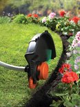 Zwarte grastrimmer met oranje spoel en wieltjes, trimt langs een bloembed.