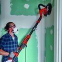Man met veiligheidsbril en masker schuurt een hoek van een muur met een lange rode schuurmachine.