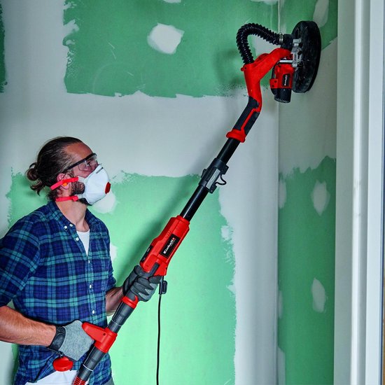 Man met veiligheidsbril en masker schuurt een hoek van een muur met een lange rode schuurmachine.