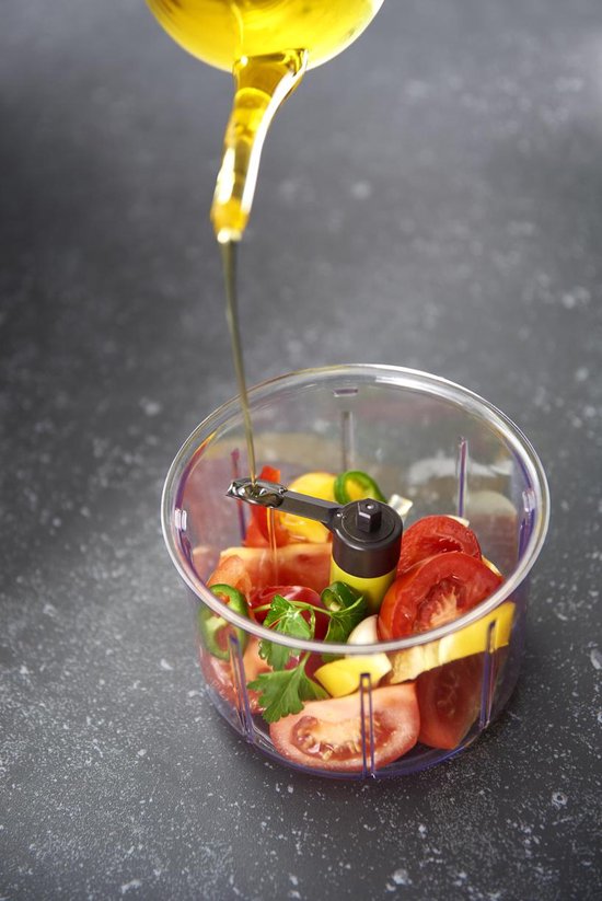 Een dunne straal olijfolie die in een transparante hakmolen met tomaten, pepers en kruiden wordt gegoten.