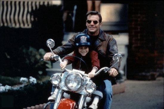 Dennis Quaid rijdt op een motor met een kind voor zich, beiden dragen helmen. Quaid draagt een leren jack.