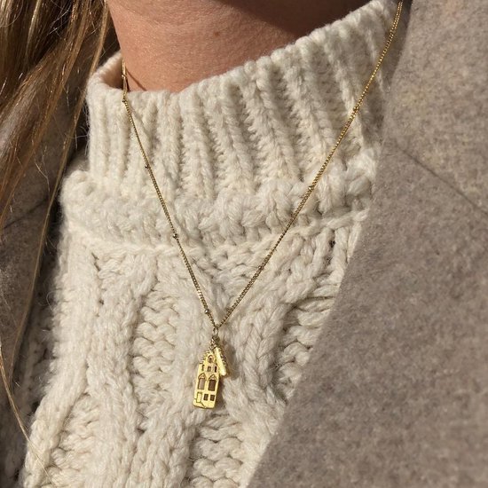 Gouden ketting met huisjeshanger op een witte gebreide coltrui.