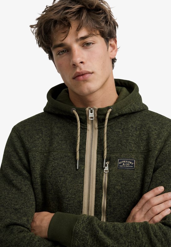 Close-up van de olijfgroene fleece hoodie met beige rits en Superdry & Co-badge op de borst.