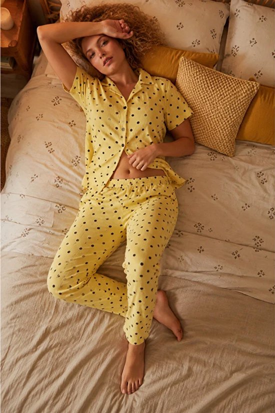 Model in een gele pyjama met korte mouwen en lange broek, bedrukt met zwarte polkadot stippen.