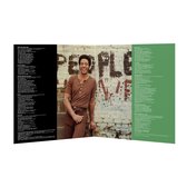 Een man, Bill Withers, staat voor een bakstenen muur waarop met zwarte verf 'PEOPLE' staat geschreven. Hij draagt een bruine coltrui en een bruine broek.