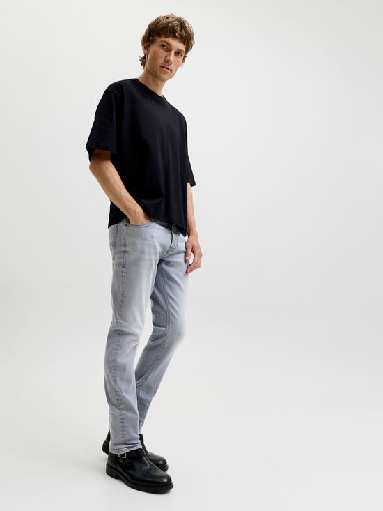 Man met hand in zak van grijze jeans, staand in de studio.