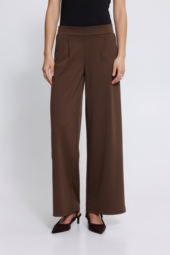 Voorkant van een model in een bruine wijde broek met plooien bij de taille, twee steekzakken en sieraden.