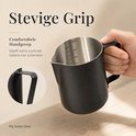 Hand pakt een zwarte RVS melkopschuimkan met een stevig handvat vast. Detail van het handvat.