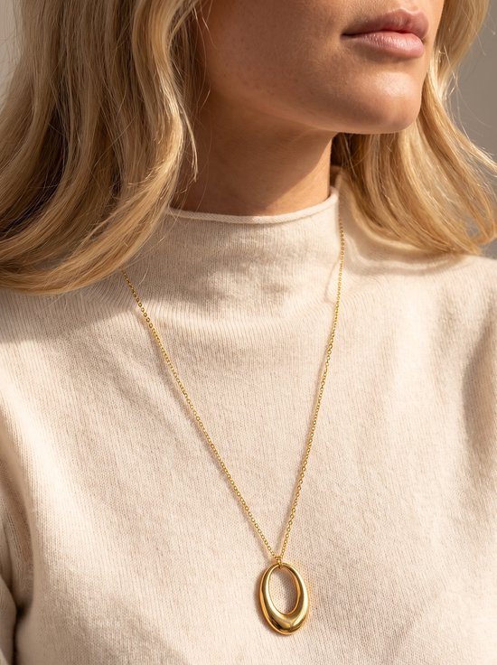 Model draagt gouden collier met ovale, holle hanger en crème coltrui.
