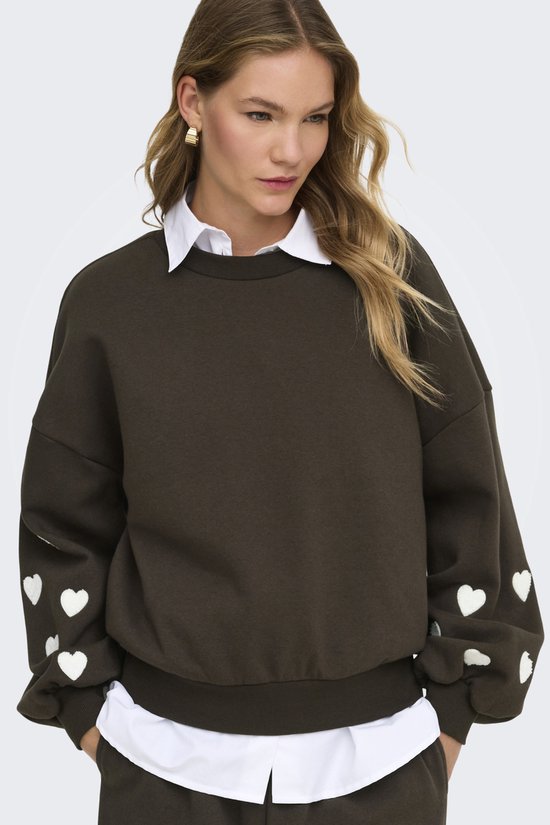 Close-up van een donkerbruine sweater met witte hartjes op de mouwen, gedragen door een model.