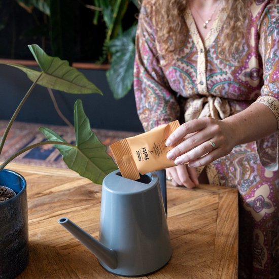 Vrouw met een kimono met paars, roze en groen patroon voegt een oranje zakje met 'Felti' toe aan een grijs gietertje.