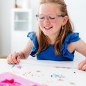 Een meisje met een bril maakt een armband met gekleurde kralen en tassels. Kralen liggen verspreid op tafel.