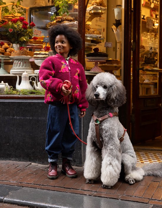 Meisje met een poedel bij een etalage vol gebak. Meisje draagt spijkerbroek en bordeaux fleece jas met poedelprint.