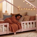 Lachend meisje ligt onder een oranje deken in een roze kinderhuisbed met decoratieve lampjes.