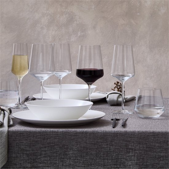 Een gedekte tafel met witte borden, witte kommen, glazen champagne, rode en witte wijn.