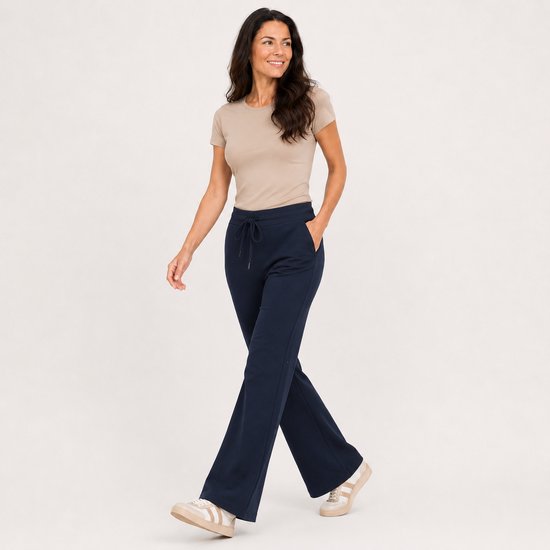 Vrouw lopend in een beige top en marineblauwe flared broek met trekkoord, handen in de zakken.