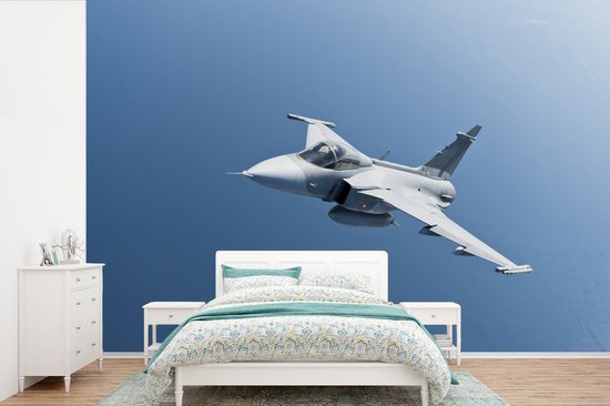 Een grijs straalvliegtuig vliegt door een blauwe hemel, afgebeeld op een muur boven een bed met patroon.