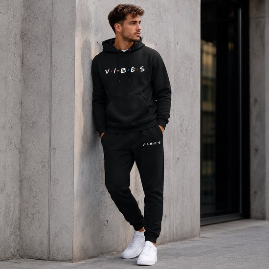 Man leunt tegen betonnen muur, draagt zwarte hoodie met 'VIBES' print en joggingbroek.