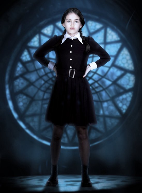 Meisje in een zwart Wednesday Addams kostuum, staand met handen in de zij voor een groot rond raam.