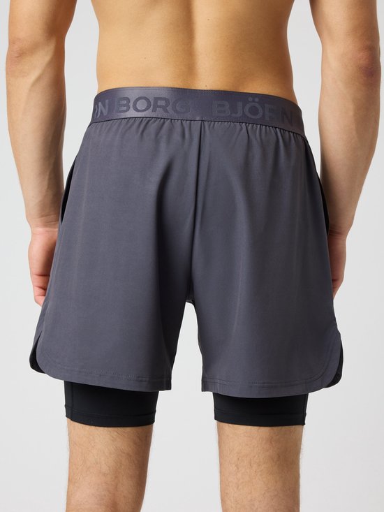 Achterkant van een donkergrijs trainingsshort met zwarte tight eronder. Logo op de tailleband.