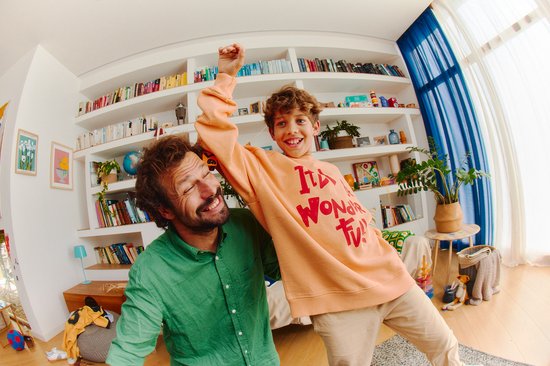 Vrolijke vader in groen overhemd en zoon met oranje trui, dansend voor een boekenkast.