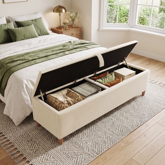 Banc de rangement blanc cassé, couvercle ouvert, rempli de couvertures et coussins, dans une chambre.
