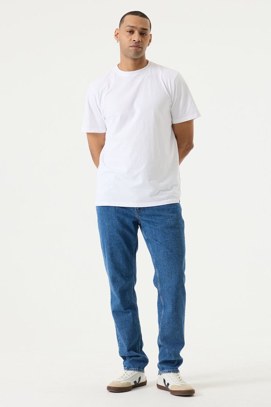Man in een witte T-shirt en blauwe jeans, staand naar voren gericht.