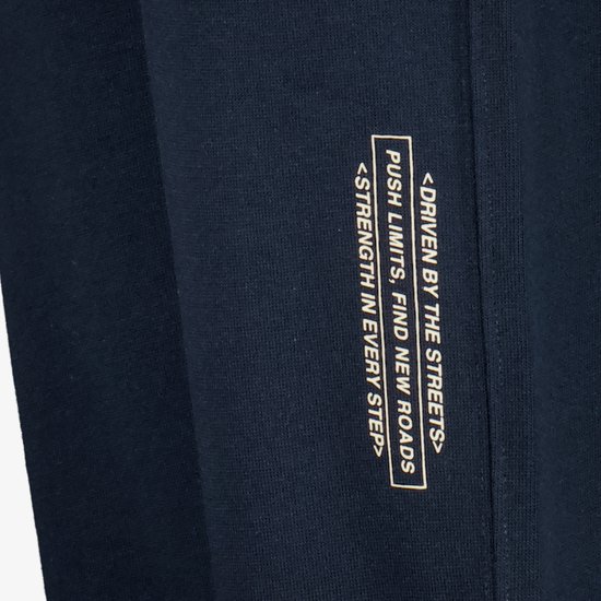 Donkerblauwe joggingbroek met gouden tekst: <DRIVEN BY THE STREETS> PUSH LIMITS, FIND NEW ROADS <STRENGTH IN EVERY STEP>
