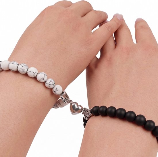 Twee handen met magnetische koppelarmbanden. Eén armband met witte stenen met grijze lijnen en zilveren details, de ander met zwarte stenen en een hartvormige magneet.