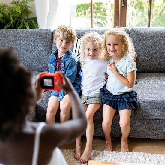 Een meisje met een rode Kidycam houdt een camera vast om een foto te maken van drie kinderen die op een grijze bank zitten.
