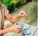 Mepal lunchpot in de kleur Nordic sage, met een grijze muesli-beker en een transparante deksel. Een persoon met een beige top en gouden armbanden schenkt muesli uit de deksel in de beker.